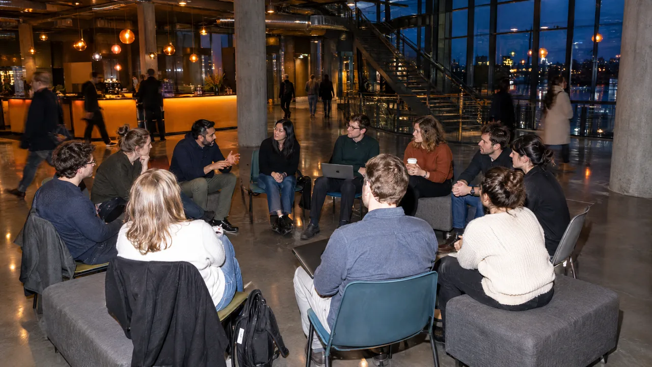 Twelve conference attendees sit in a loose circle of chairs and benches in an evening conference foyer, one speaker gesturing while the others listen