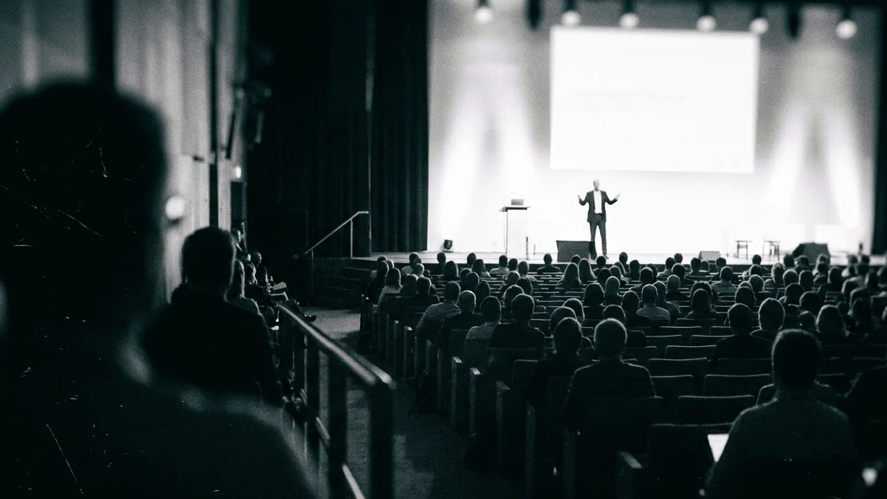 Delegates leaving a conference session room between talks, small groups standing near rows of chairs while a technician adjusts a microphone on the stage