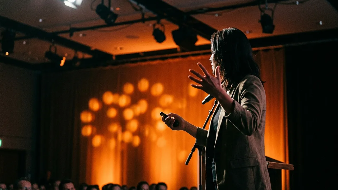 A speaker with shoulder length dark hair and a brown blazer stands in half profile at the edge of a conference stage, one hand raised in an open gesture and a presentation clicker in the other, behind a microphone stand, with a warm orange light pattern on the curtain behind her and the heads of the audience in the dark at the lower edge