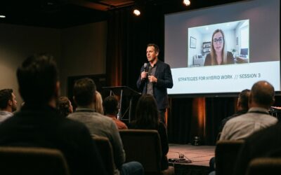 Speaker on stage at a mid-sized business conference with a remote participant displayed on the large screen behind the speaker; audience heads softly out of focus in the foreground.